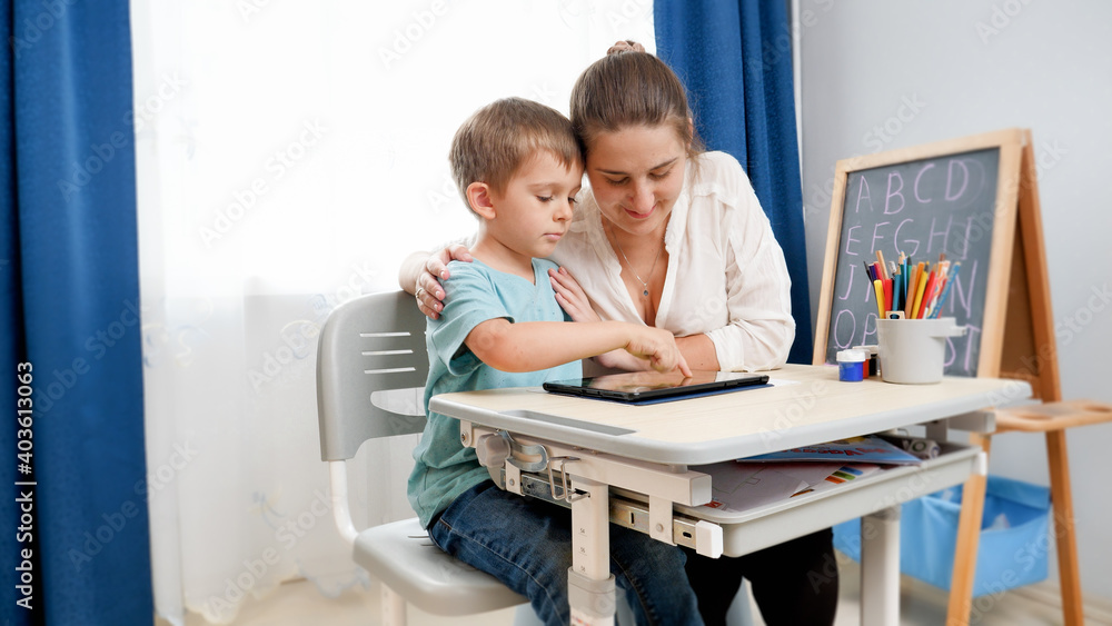 Young mother hugging her little son doing homework and studying on tablet computer at home. CHild using gadget for education and learning Young mother hugging her little son doing homework and studying on tablet computer at home. CHild using gadget for education and learning