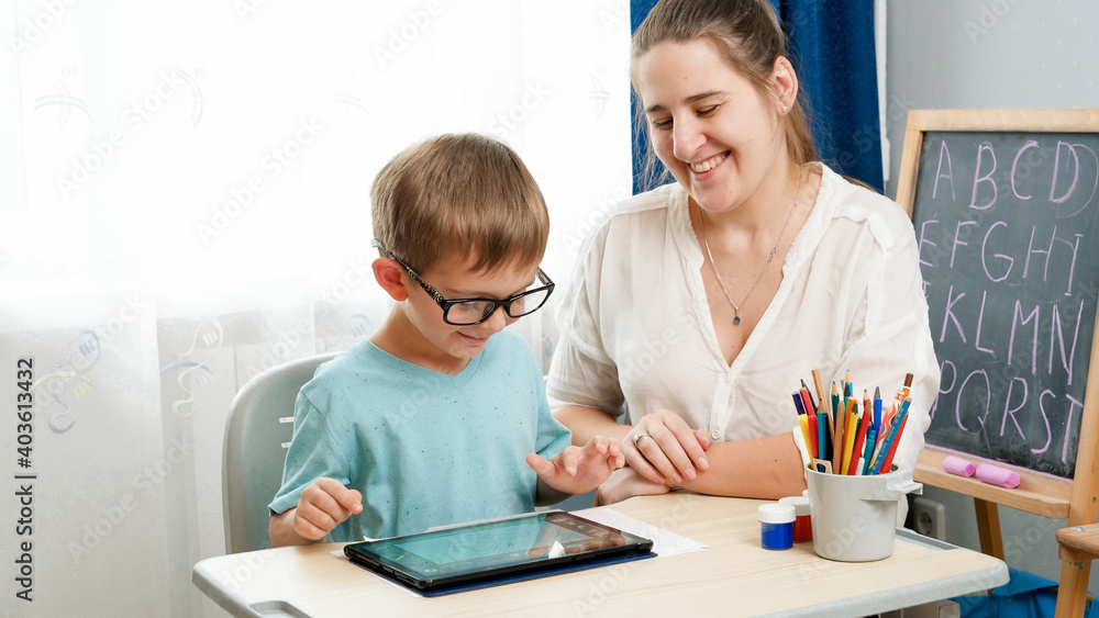 Little boy in eyeglasses playing on tablet computer. Mother hugging her little son doing homework using gadet. Children having problems with eyes and sight Little boy in eyeglasses playing on tablet computer. Mother hugging her little son doing homework using gadet. Children having problems with eyes and sight