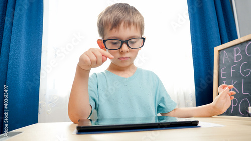 Portrait of smart cute boy wearing glasses playing and studying on tablet computer. CHild using gadget for doing homework