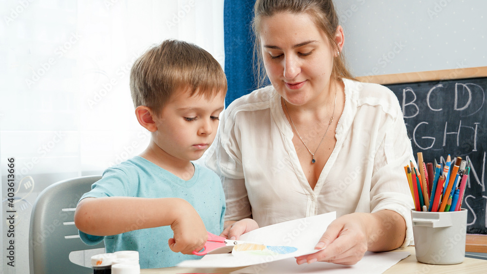 Young female teacher showing her little student how to use scissors. Boy cutting paper with scissors while doing homework. Young female teacher showing her little student how to use scissors. Boy cutting paper with scissors while doing homework.