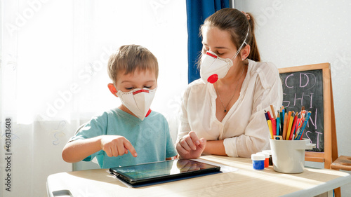 Little boy wearing protective medical mask respirator studying in classroom with female teacher. Doing homework and studying at home with parents during lockdown and self isolation.