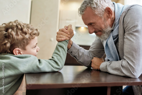 Enjoying time at home. Happy grandfather and little grandson sitting opposite each other and playing arm wrestling, having fun together