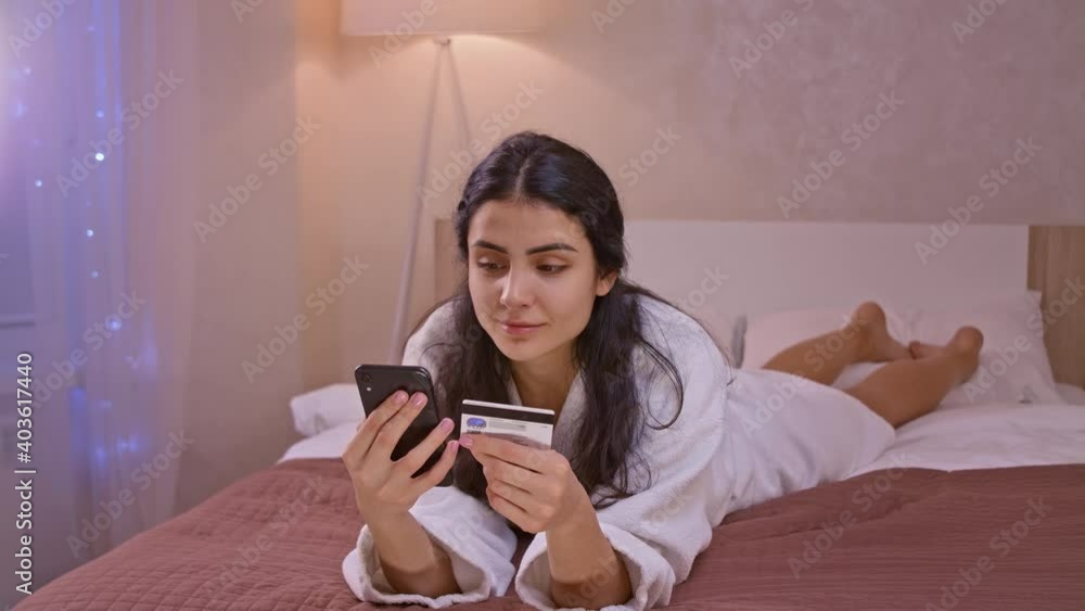 Smiling young woman customer holding credit card and smartphone sitting on couch at home. Happy female shopper using instant easy mobile payments making purchase in online store. E-banking app service Smiling young woman customer holding credit card and smartphone sitting on couch at home. Happy female shopper using instant easy mobile payments making purchase in online store. E-banking app service