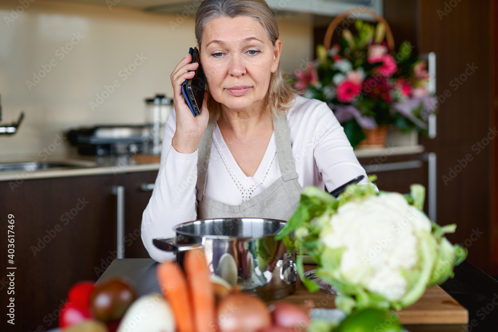 Senior woman in kitchen preparing food and holding smart phone.