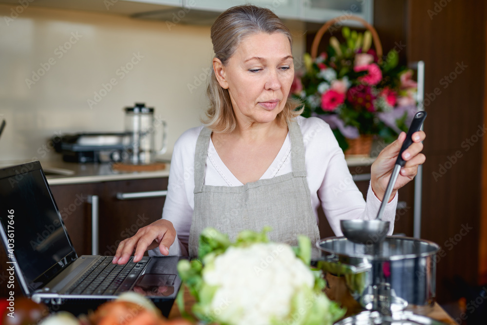 Senior woman in apron at kitchen prepares food and uses laptop.
