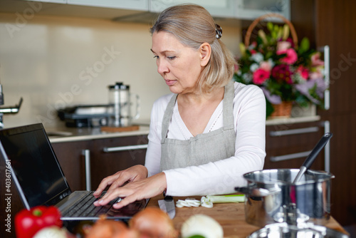 Mature woman in kitchen prepares food and looks into laptop.