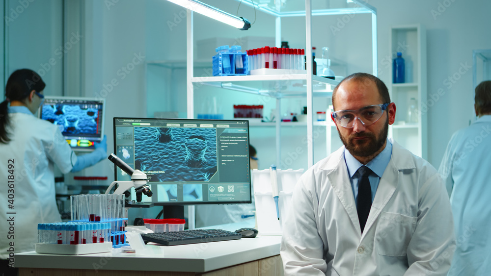 Portrait of exhausted chemist looking at camera in modern equipped lab late at night. Team of biochemists examining virus evolution using high tech and chemistry tool for research, vaccine development Portrait of exhausted chemist looking at camera in modern equipped lab late at night. Team of biochemists examining virus evolution using high tech and chemistry tool for research, vaccine development