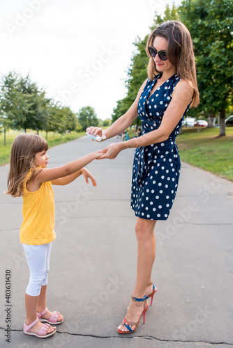 Woman mom with her daughter girl, antiseptic from viruses of diseases and microbes, treatment of hands and skin from viruses, dirt and wounds on hand. Summer casual clothes in park.