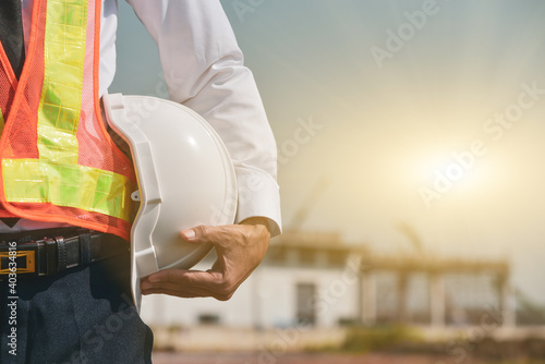Construction foreman holding white hat safety hard hat on building background