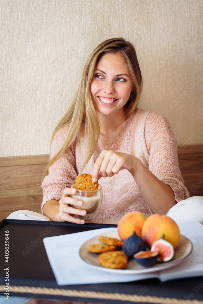 Efficient young millennial attractive woman sitting on a bed in the morning. Hand holding a cup of coffee and eating cookies. Coffee morning concept. Pleasant time during calm morning Efficient young millennial attractive woman sitting on a bed in the morning. Hand holding a cup of coffee and eating cookies. Coffee morning concept. Pleasant time during calm morning