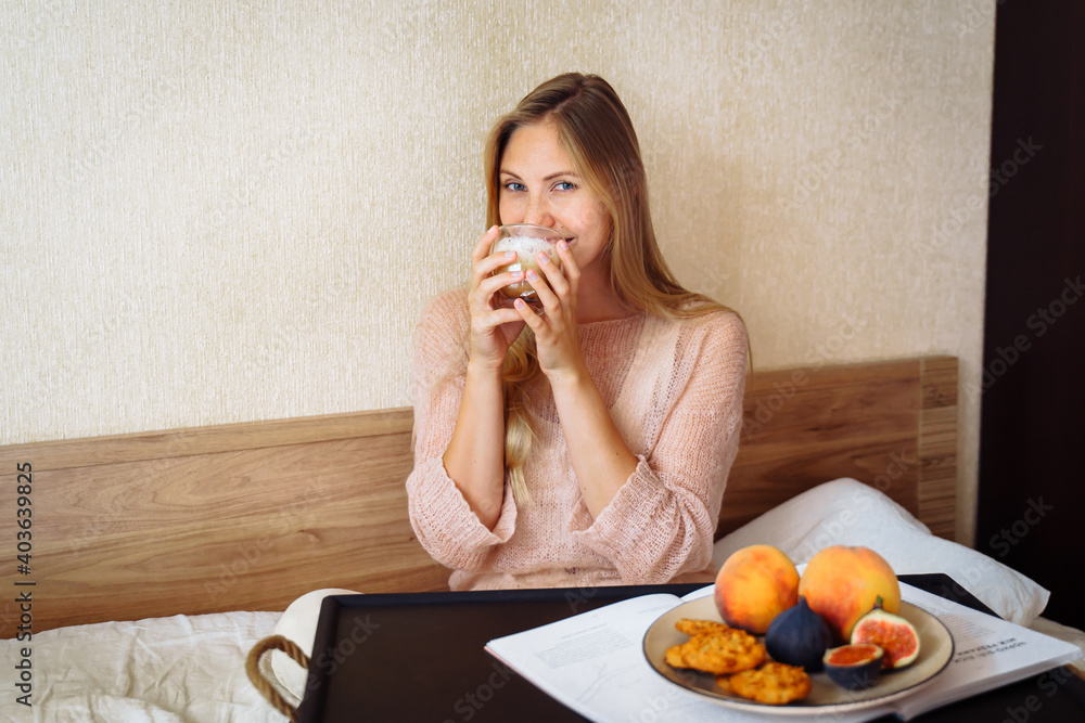 Good morning. Attractive girl drinking morning coffee in bed. Sitting on snow-white bed in the bedroom, in front of her stands for breakfast with various bright fruits and pastries. Pleasant and calm Good morning. Attractive girl drinking morning coffee in bed. Sitting on snow-white bed in the bedroom, in front of her stands for breakfast with various bright fruits and pastries. Pleasant and calm