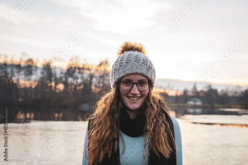 Lächelnde junge Frau mit einer Wollmütze und Brille an einem See. Portrait, langes lockiges Haar, Winter. photo