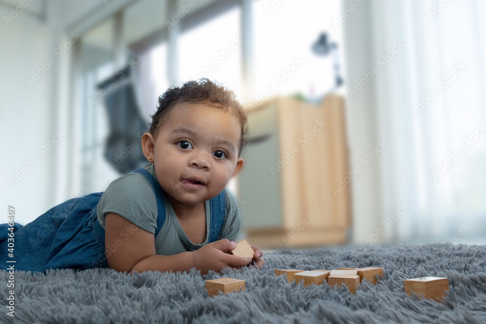 Fototapeta premium Little dark skinned girl lay on the carpet at home, and playing wooden toys at home, looking at camera