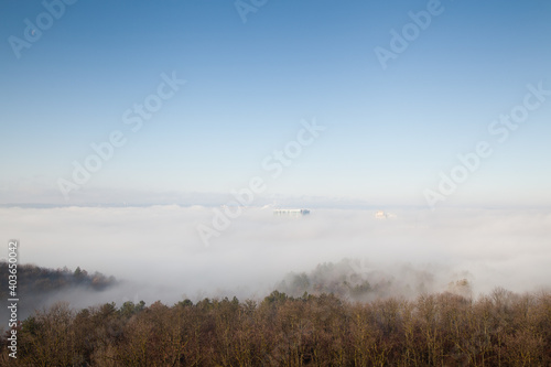 Trees with roofs of several buildings in the fog with blue sky