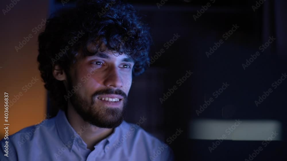 Close-up face of smiling young man watching on monitor screen and working on computer at home office workplace at late night in dark room, tracking shot. Concept of remote working, distance learning. Close-up face of smiling young man watching on monitor screen and working on computer at home office workplace at late night in dark room, tracking shot. Concept of remote working, distance learning.