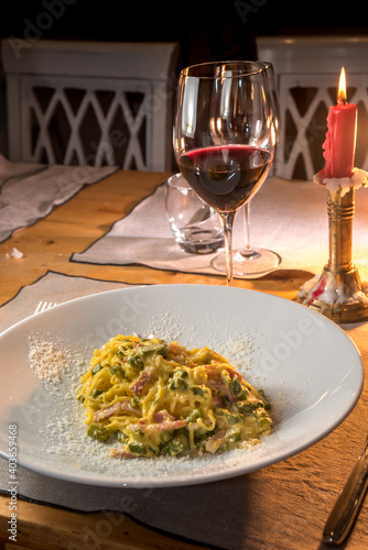 Carbonara fettuccine with asparagus. Egg spaghetti with bacon, eggs and grated parmesan in plate on the set table of trattoria with glass of wine and red candle lit