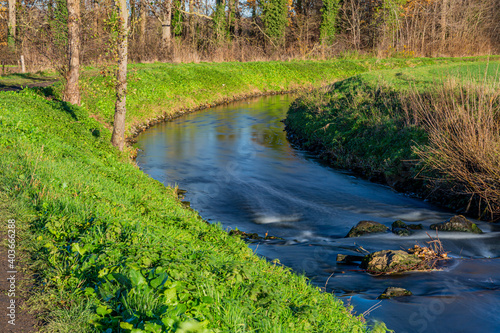 Small winding river with a calm flow between stones among green grass and trees, sunny winter day in Sweikhuizen, South Limburg, the Netherlands. Long exposure photography photo