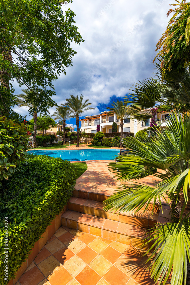 Fototapeta premium Swimming pool, sun-loungers and palm trees during a warm sunny day, paradise destination for vacations. Backyard swimming pool with garden full of palm trees and flowers. Backyard with swimming pool.