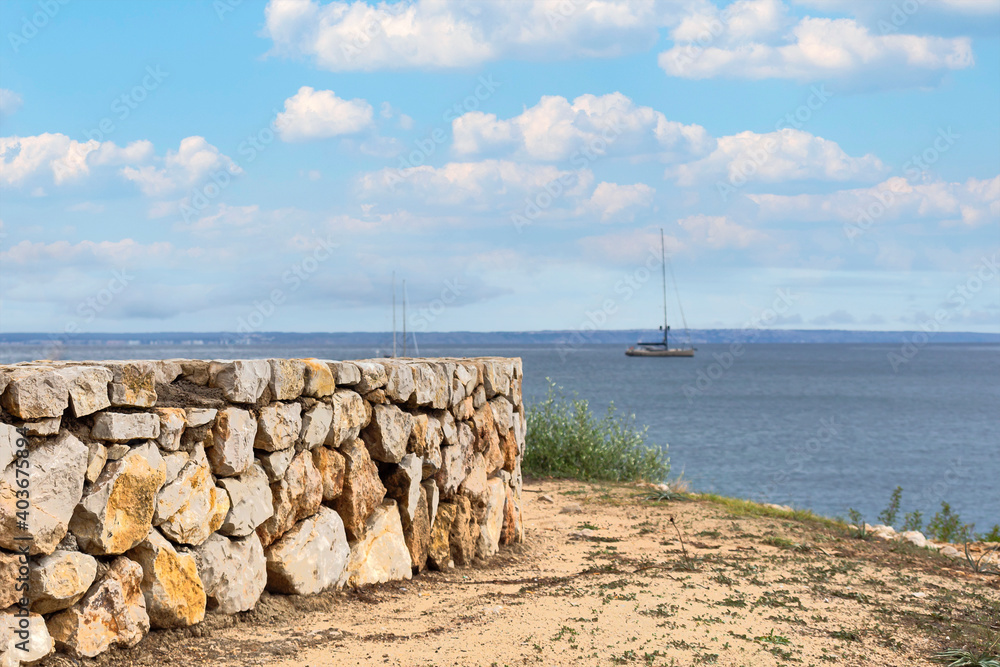 view from the sandy seashore with a stone wall to the blue sea with a yacht on the horizon