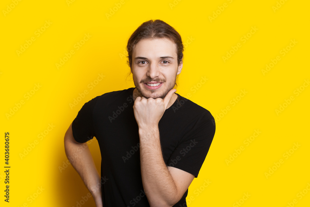 Handsome bearded man with long hair is touching her chin with the fist and smile at camera on a yellow studio wall Handsome bearded man with long hair is touching her chin with the fist and smile at camera on a yellow studio wall