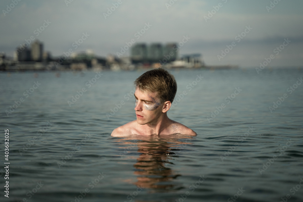 portrait of a non-binary young man with sparkles on his face, standing in the sea, in the background you can see the city