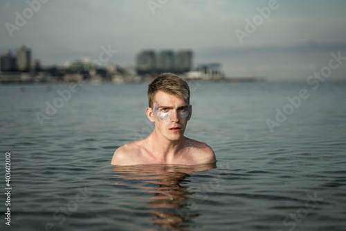 portrait of a non-binary young man with sparkles on his face, standing in the sea, in the background you can see the city © Maxim