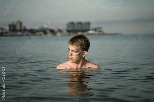 portrait of a non-binary young man with sparkles on his face  standing in the sea  in the background you can see the city