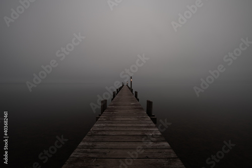 Mole (pier) on the „Chiemsee“ lake in Bavaria, Germany. Wooden bridge in winter time with frozen and icy lake and dim lanterns for fishing. Dark and Foggy lake.