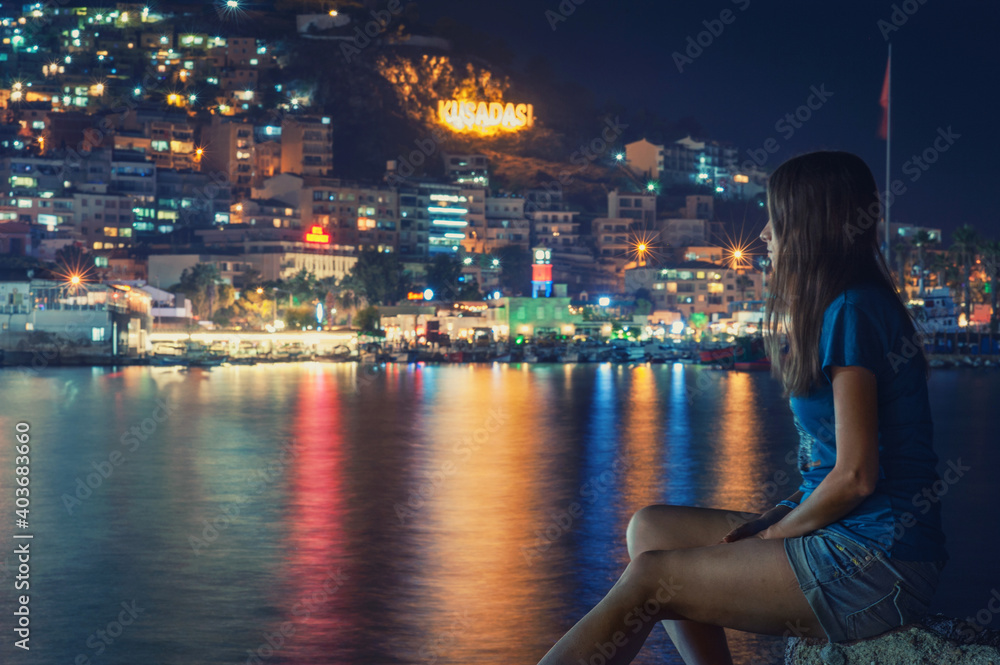Young woman is sitting on the rocks by the sea at night in Kusadasii, Turkey Young woman is sitting on the rocks by the sea at night in Kusadasii, Turkey