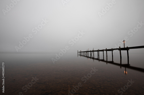 Mole  pier  on the    Chiemsee    lake in Bavaria  Germany. Wooden bridge in winter time with frozen and icy lake and dim lanterns for fishing. Dark and Foggy lake.  