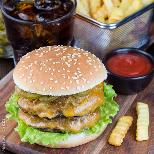 Double cheese burger with caramelized onion, lettuce and jalapeno, served with french fries and soda, on a wooden board, square, closeup