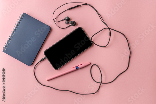 notepad, pen and smartphone with headphones on a pink surface, top view, mock up