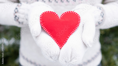 A red heart as a gift in the hands of a woman. Love declaration concept. A girl in a cozy white knitted sweater and mittens holds out a heart on a green blurred background. Cute valentine card