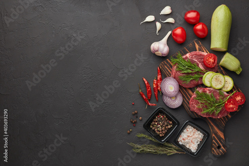Beef steaks on cutting board, vegetables and spices on black slate background. Top view. Steak menu