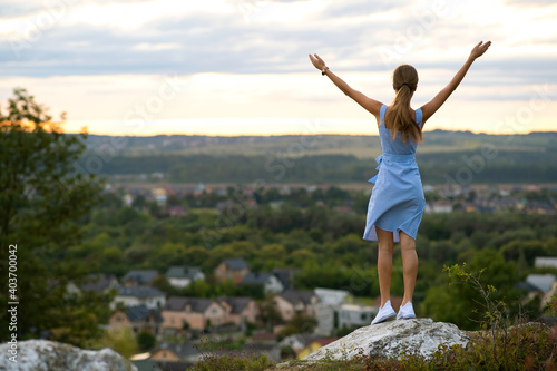 A young woman in summer dress raising up her hands standing outdoors enjoying view of bright yellow sunset.