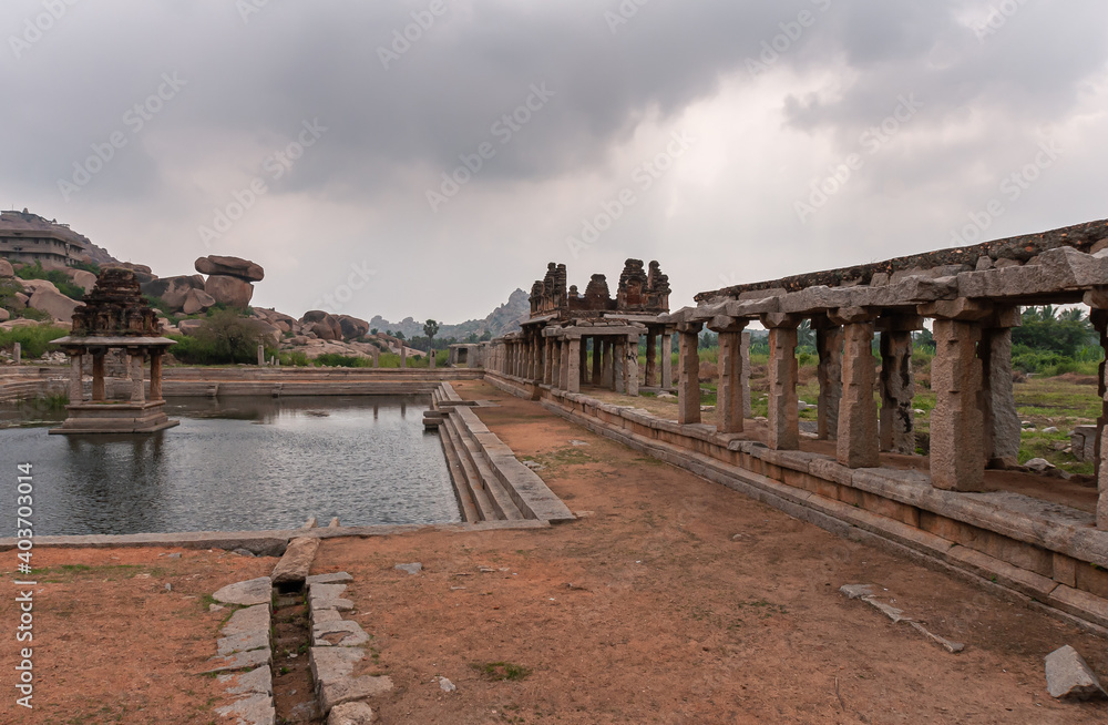 Hampi, Karnataka, India - November 5, 2013: Sri Krishna tank in ruins. south side of pool with shrine and gallery on the side under gray cloudscape. Boulders and mountain on horizon.