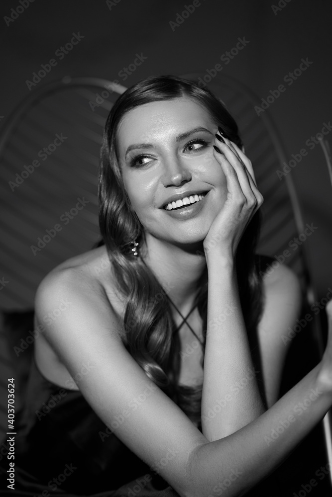Fototapeta premium Elegant woman with perfect makeup and wavy brunette hair in long black silk evening dress sitting at sofa in luxury stylish interior