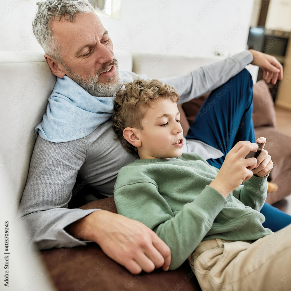 Children and gadgets. Cute little boy playing video game on smartphone while relaxing on sofa with his happy grandfather Children and gadgets. Cute little boy playing video game on smartphone while relaxing on sofa with his happy grandfather