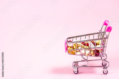 Shopping cart with different medical pills and capsules in it on pink background. Pharmacology and drugstore concept.