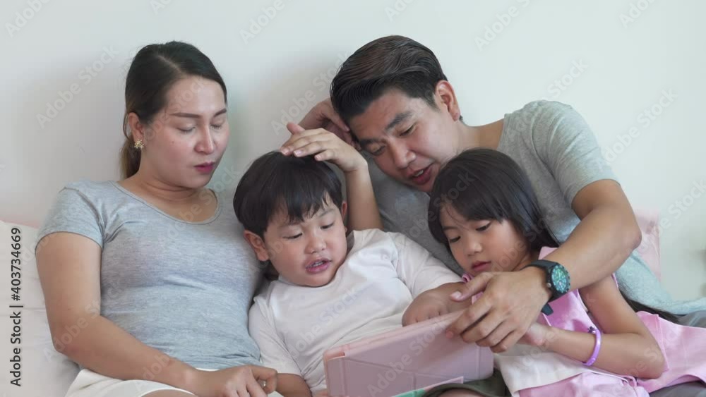 Happy Asian family parents with little daughter and son sitting on bed and using digital tablet with internet together. Father, mother and kids relax and having fun with wireless technology at home. Happy Asian family parents with little daughter and son sitting on bed and using digital tablet with internet together. Father, mother and kids relax and having fun with wireless technology at home.