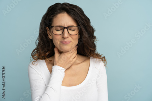Sick woman having sore throat, tonsillitis, feeling sick, suffering from painful swallowing, angina, strong pain in throat, loss of voice, holding hand on her neck, isolated on studio blue background.