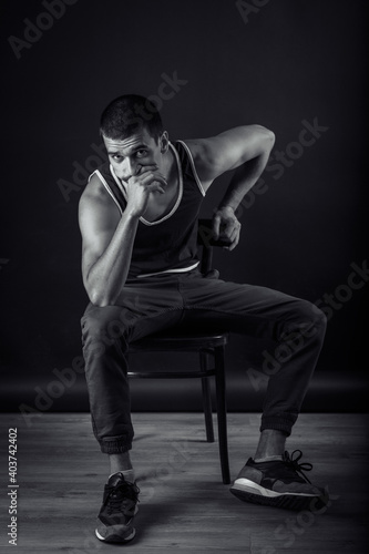 black and white handsome tanned sports man in a T-shirt sits thoughtfully on a chair on a black background