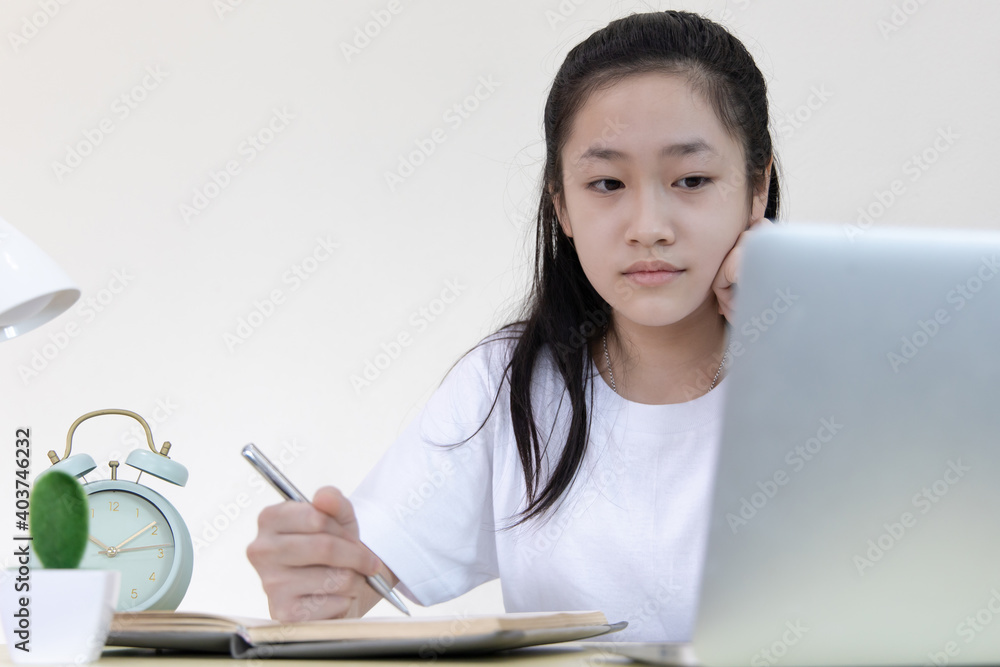 Asian girl is studying on a laptop and writes in a notebook with intention,Online learning, stay home, New normal, Covid-19 coronavirus, Social distancing, Internet learning...