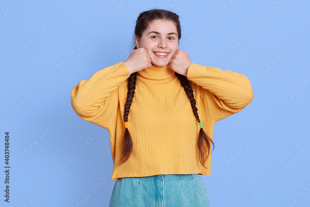 Portrait of pretty smiling young woman in casual yellow sweater and jeans clenching fists near cheeks isolated on pastel blue background, having sincere emotions, has dark hair and two pigtails. Portrait of pretty smiling young woman in casual yellow sweater and jeans clenching fists near cheeks isolated on pastel blue background, having sincere emotions, has dark hair and two pigtails.