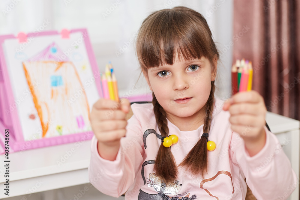 Little girl learning to draw, holding in both hands many colored wax crayons and pencils, kid looking at camera and showing her equipment for painting. Little girl learning to draw, holding in both hands many colored wax crayons and pencils, kid looking at camera and showing her equipment for painting.