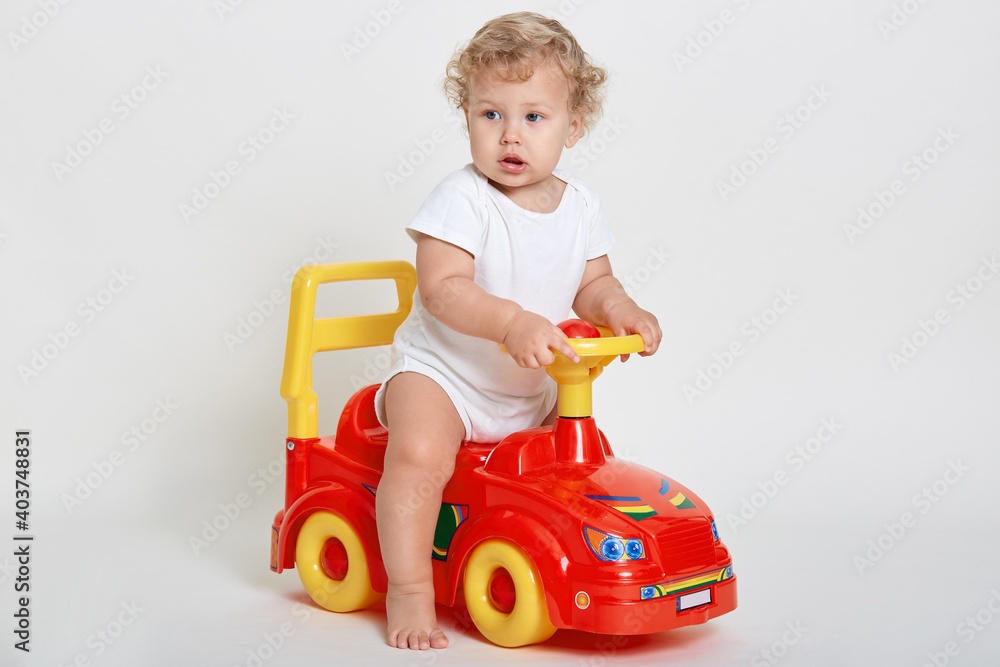 Curious child sitting in toy car and looking away, sees something interesting away, having curious facial expression, posing barefoot isolated over white background. Curious child sitting in toy car and looking away, sees something interesting away, having curious facial expression, posing barefoot isolated over white background.