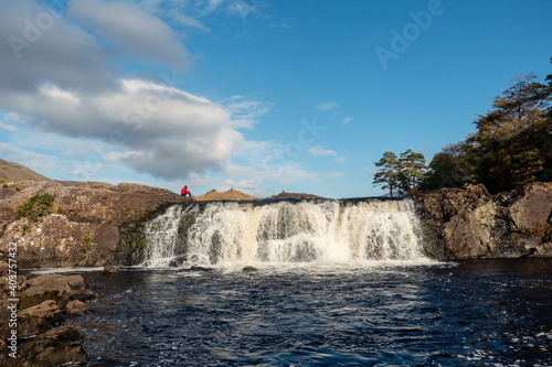Aasleagh waterfall in county Mayo, Ireland. Warm sunny day, blue sky and water. Popular tourist attraction. Teenager in a red hoodie sitting and looking at running water.