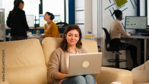 Young woman entrepreneur sitting on couch in middle of business start up office looking at camera smiling holding laptop. Multiethnic coworkers talking about startup financial company in modern office © DC Studio