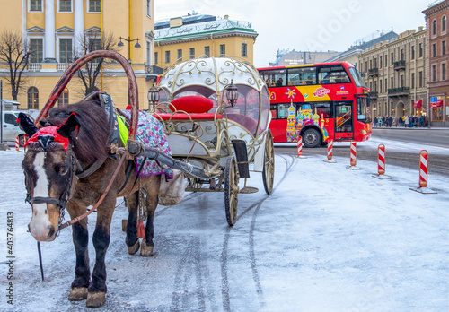 In winter, brown horse harnessed to a historic carriage stands on a city street with a tourist bus photo