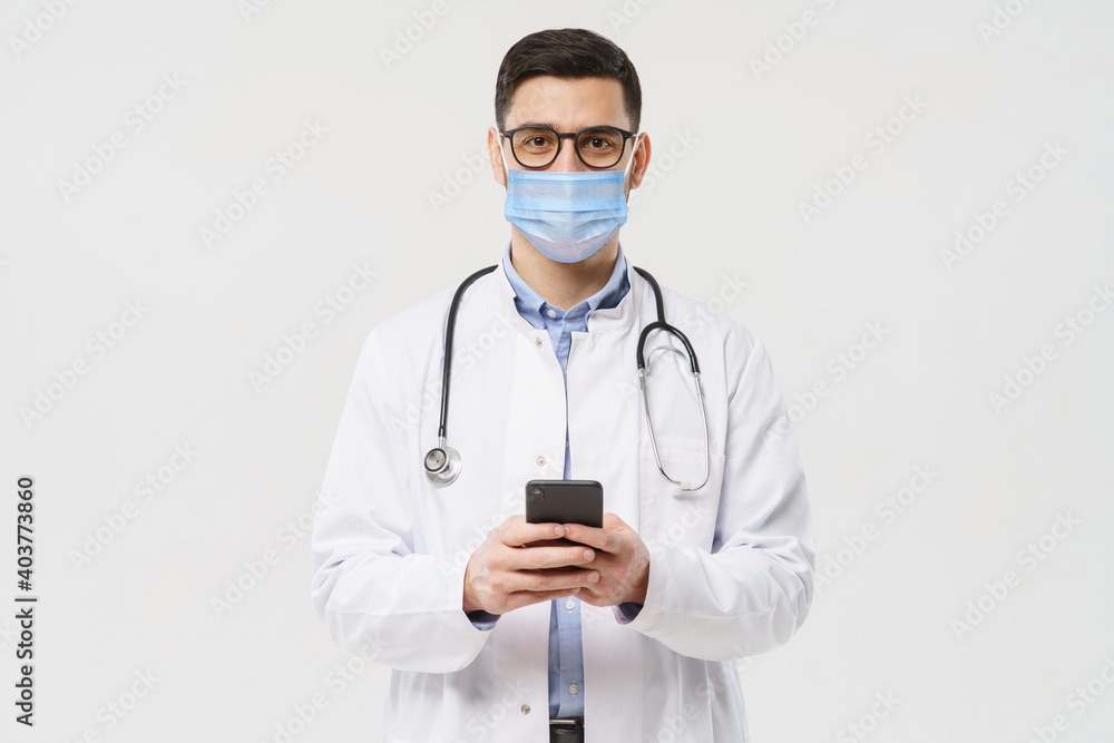 Portait of young doctor wearing surgical mask and white coat, holding his phone with both hands, using medical app, isolated on gray background Portait of young doctor wearing surgical mask and white coat, holding his phone with both hands, using medical app, isolated on gray background
